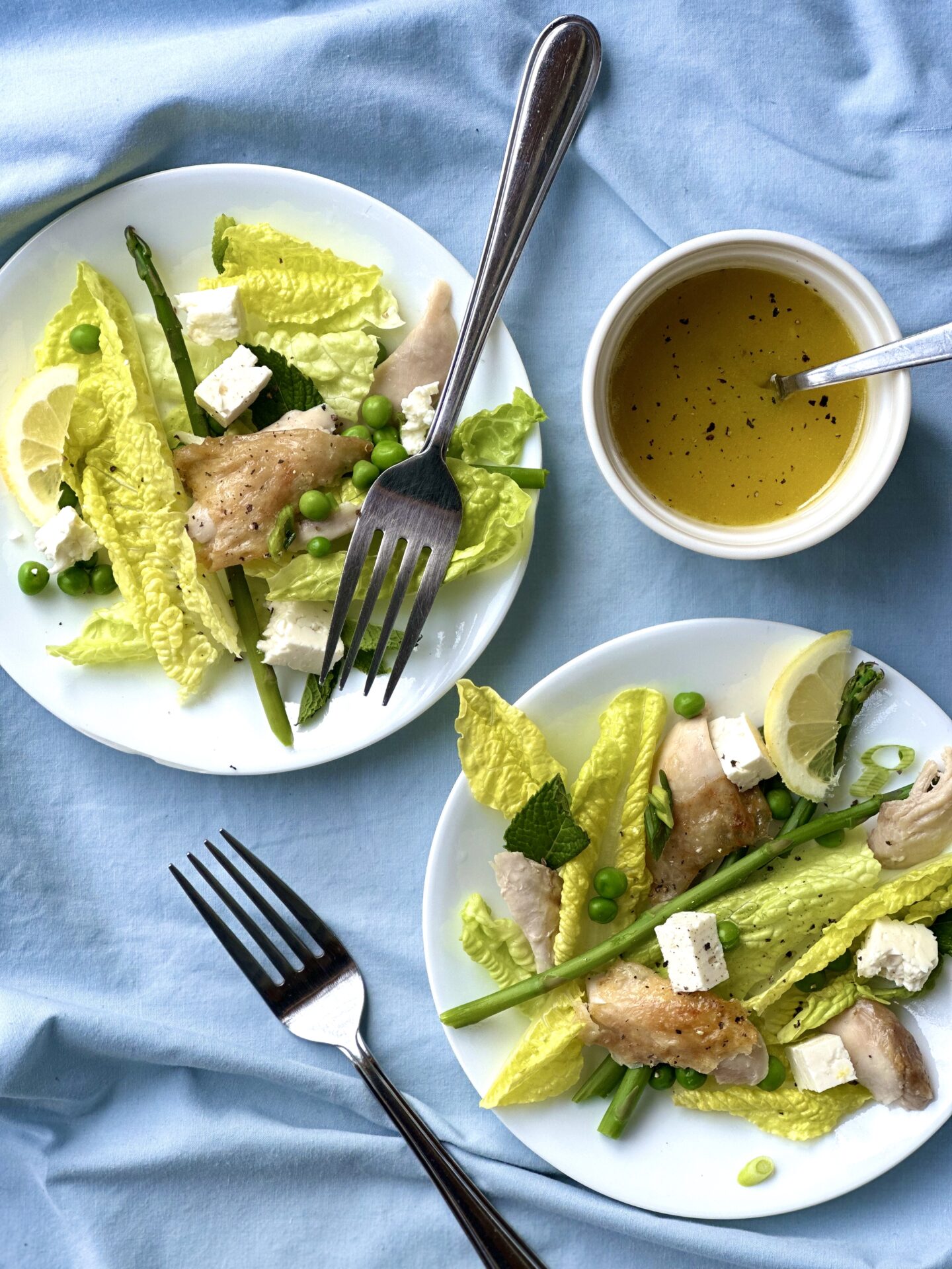 078EDF63-95AD-43B9-A73B-62F8C748961C Two plates of salad with romaine lettuce, roast chicken and lemon seen with a bowl of dressing on a blue back ground