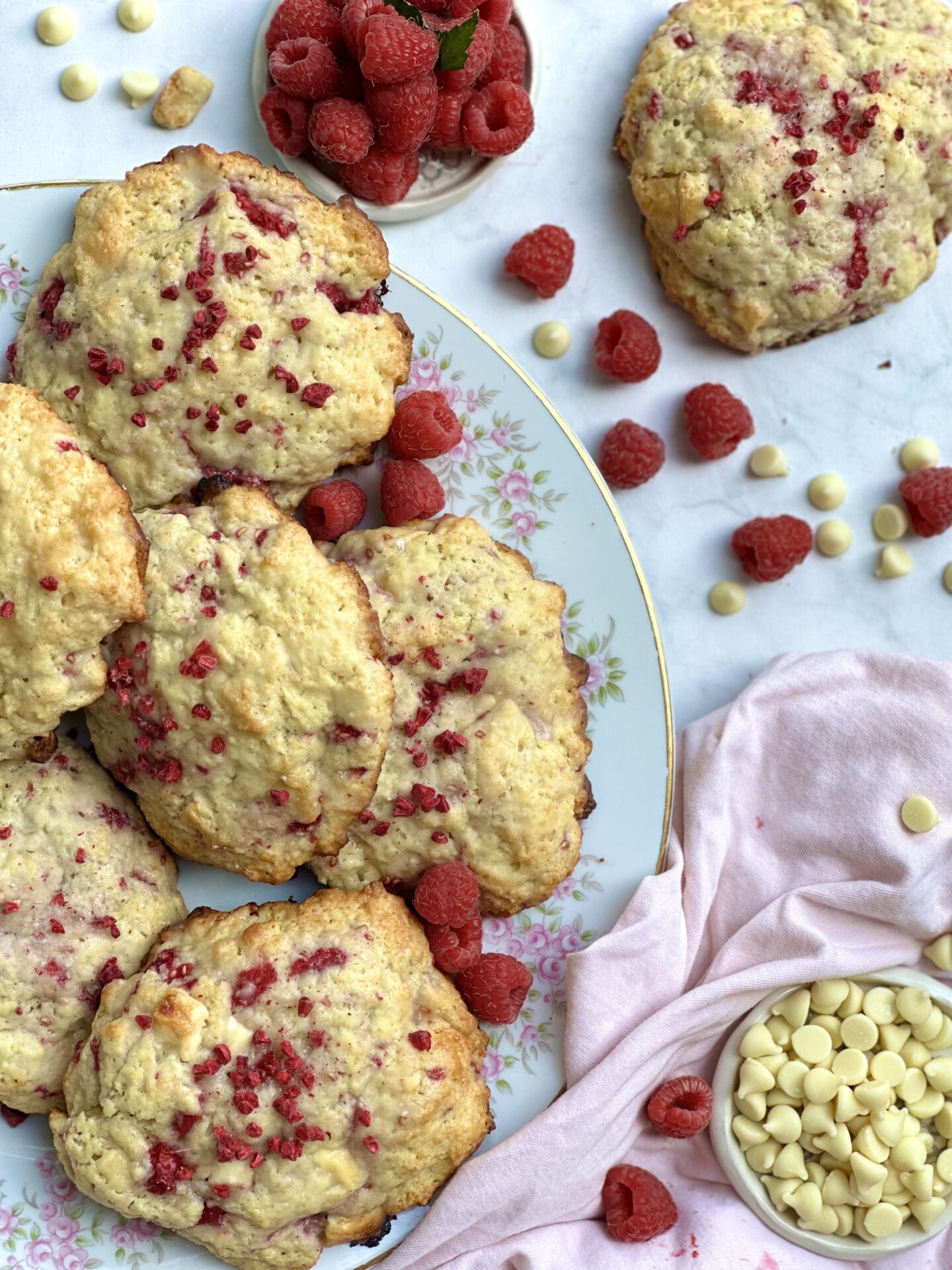 Vintage china plate with pink rosebuds piled high with freshly baked Raspberry White Chocolate Scones, surrounded by fresh red raspberries and white chocolate chips
