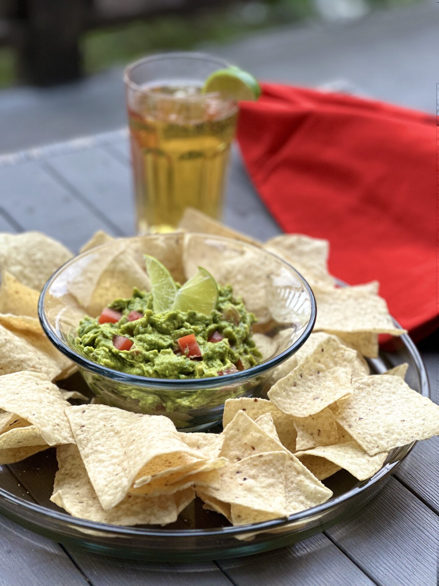 A platter of chips with fresh homemade guacamole sitting on an outdoor patio table with a glass of cold beer and a bright red napkin