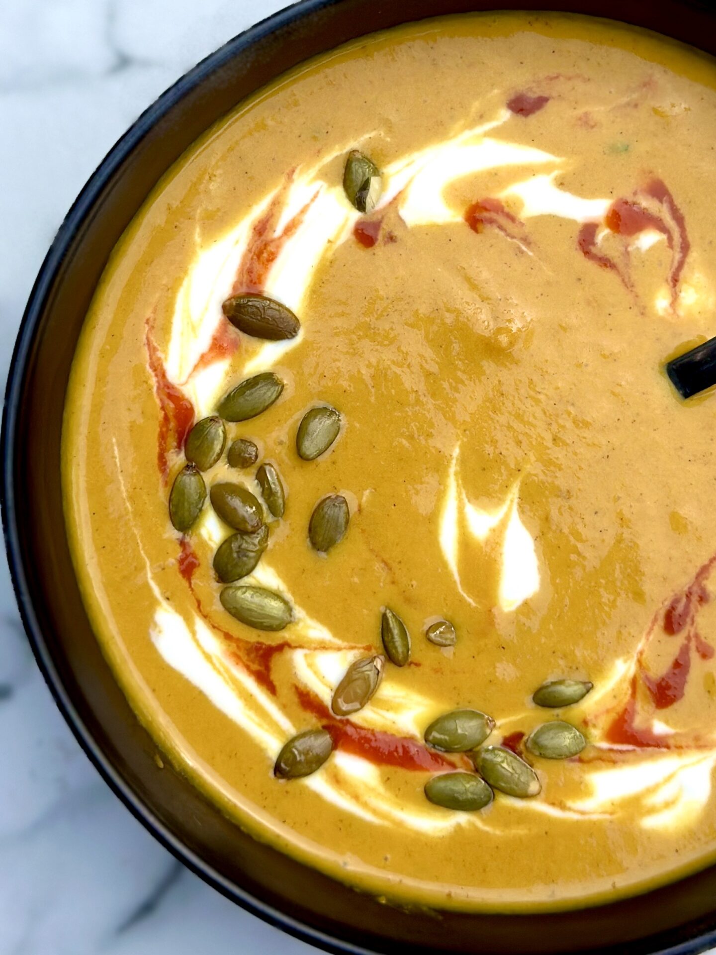 A black bowl filled with golden yellow Creamy Pumpkin Spice Soup is seen from above, garnished with swirls of white yogurt and red sriracha, and sprinkled with pumpkin seeds