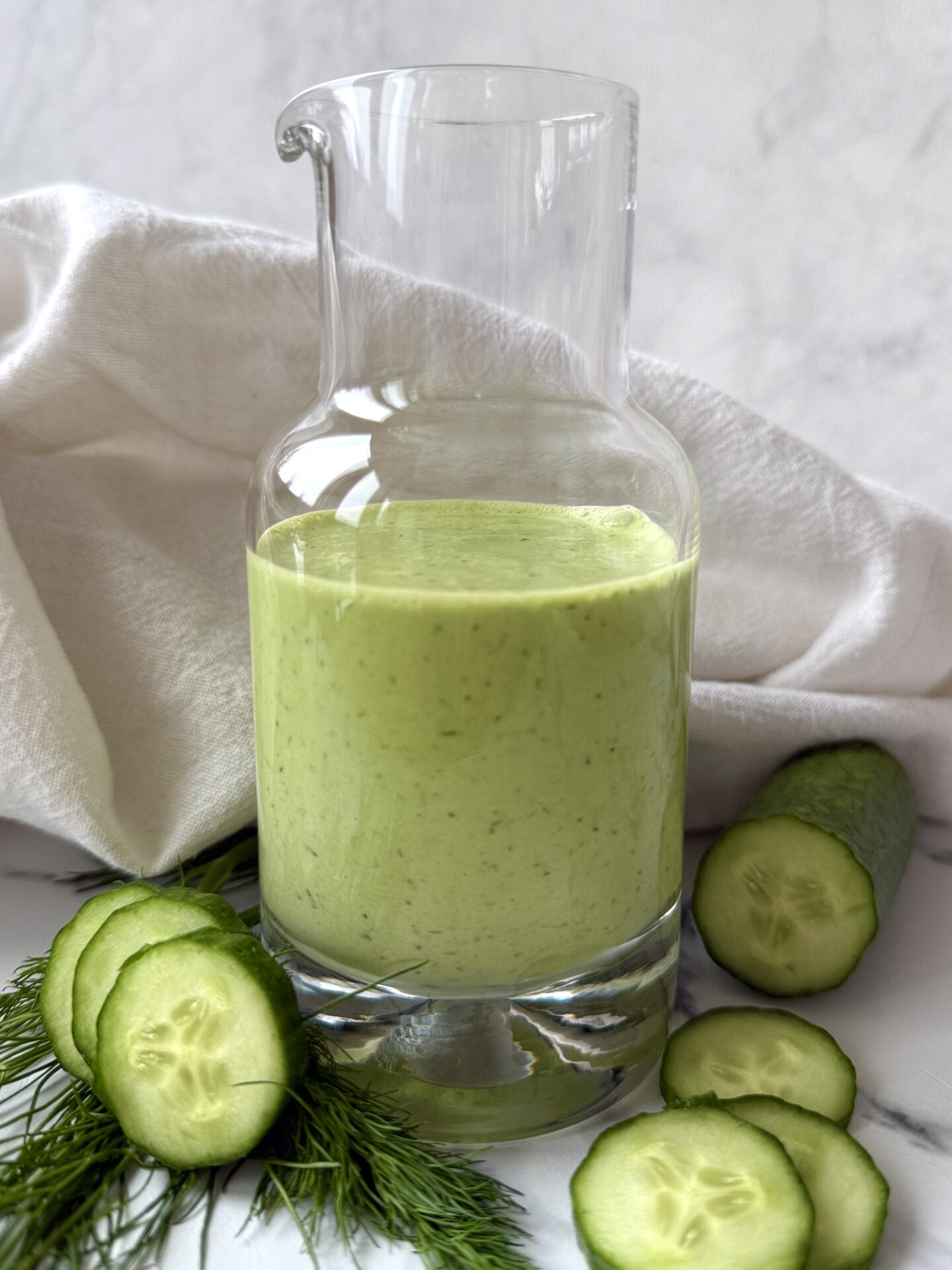 A glass bottle of Creamy Cucumber Dill Salad Dressing is seen on a white background, surrounded by fresh dill and sliced cucumbers