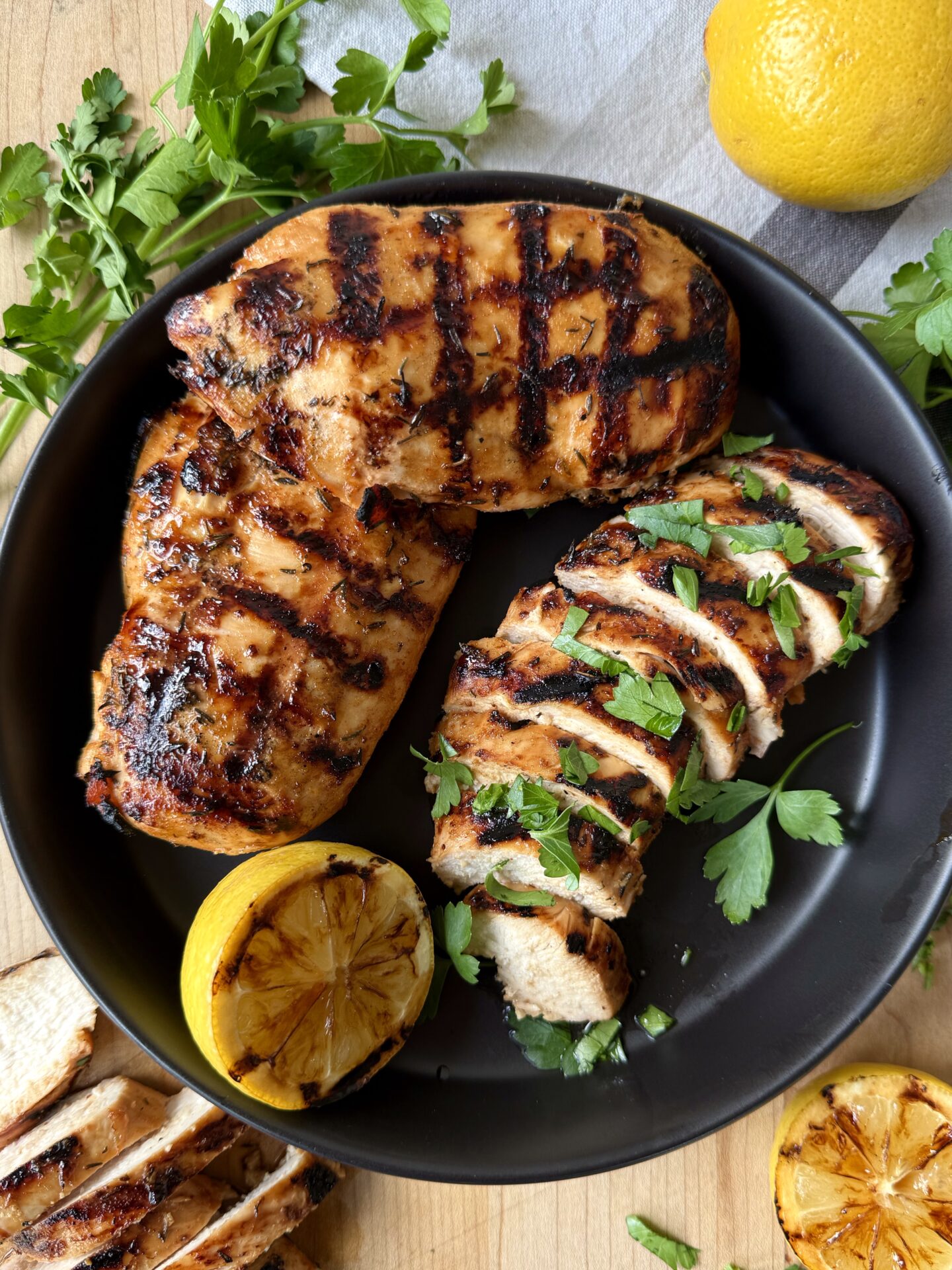 A black platter is seen from above, filled with Sweet and Tangy Marinated Grilled Chicken.  One chicken breast is sliced, whereas the others are seen whole, with perfect crosshatch grill marks.  The chicken is garnished with grilled lemon halves and fresh parsley.