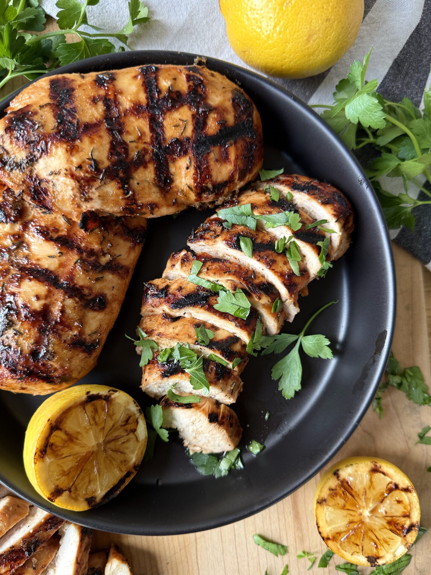 A black plate overflows with sweet and tangy marinated grilled chicken. Grilled lemons and fresh parsley garnish the dish.