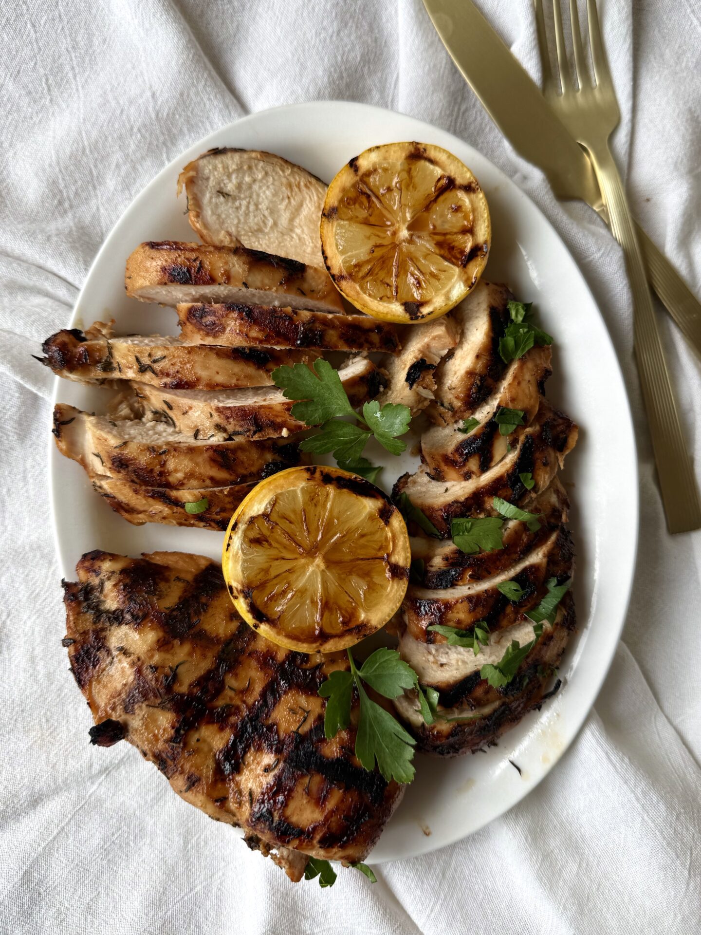 A white platter is seen from above, filled with Sweet and Tangy Marinated Grilled Chicken.  Some of the chicken breasts are sliced, whereas others are seen whole displaying their crosshatched grill marks.  The platter rests on a white linen tablecloth.  Gold coloured utensils are seen to the side.