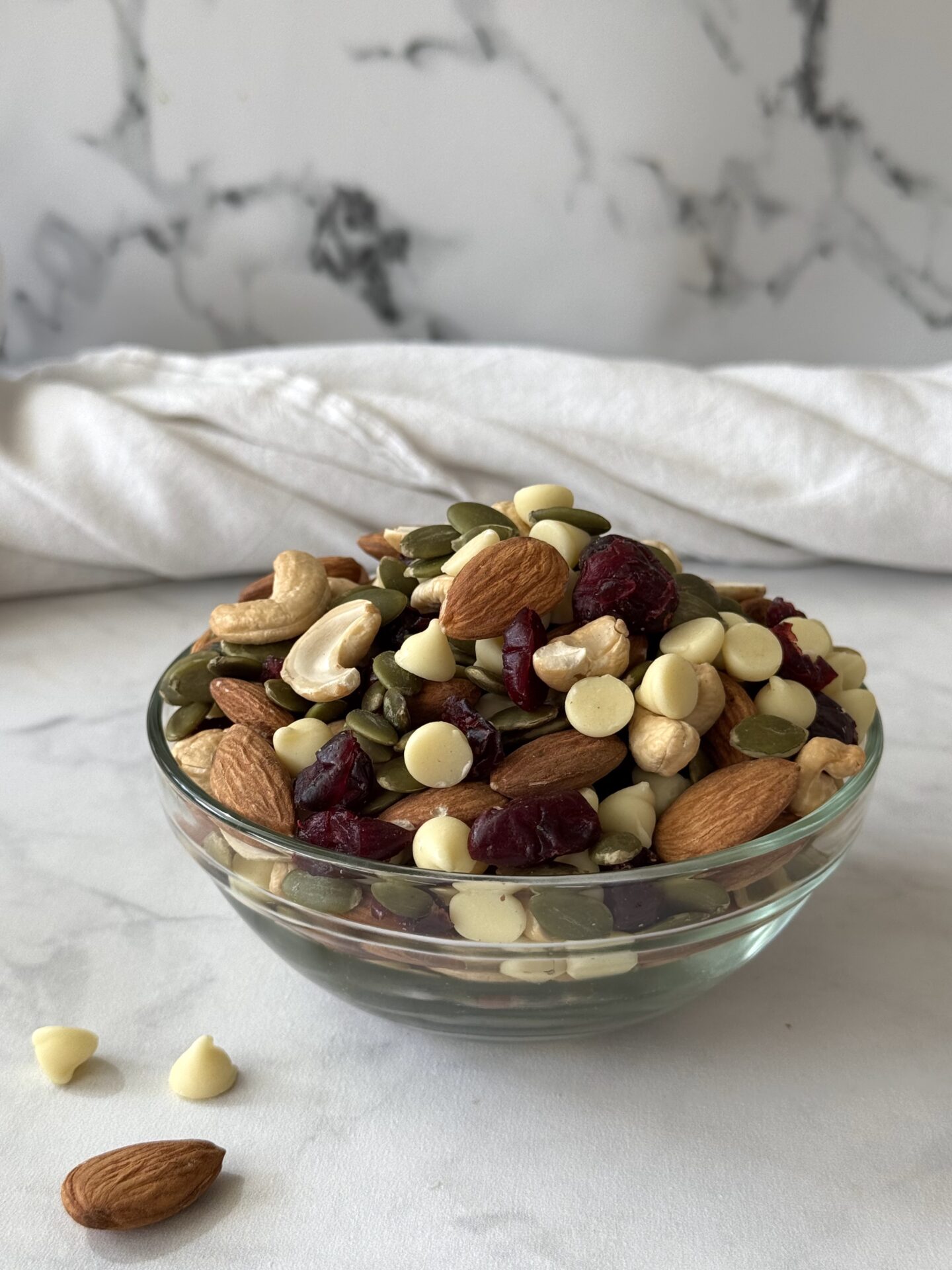 A glass bowl of White Chocolate and Cranberry Trail Mix is seen on a white marble countertop. A few white chocolate chips and a whole almond sit beside the bowl.
