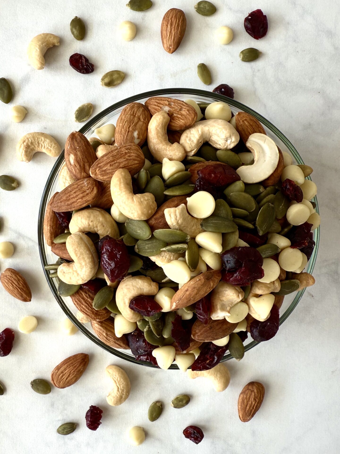 A bowl of White Chocolate and Cranberry Trail Mix is seen from above. Scattered almonds, cashews, pumpkin seeds, dried cranberries and white chocolate chips can be seen scattered on the table around the bowl, on the left side.