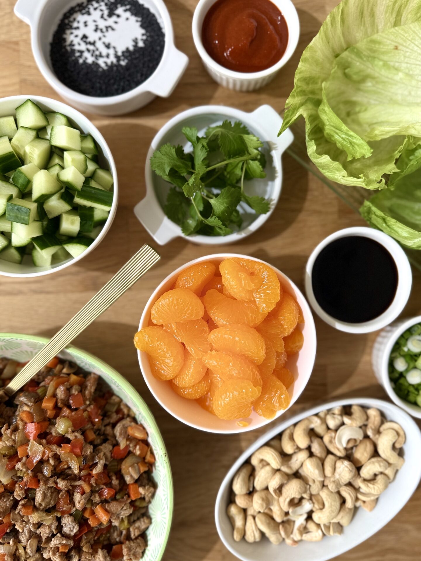Bowls containing the toppings for turkey lettuce wraps are seen seen from above. These bowls offer a colourful array of Mandarin orange slices, chopped cucumber, cashew nuts, sesame seeds and sauces.