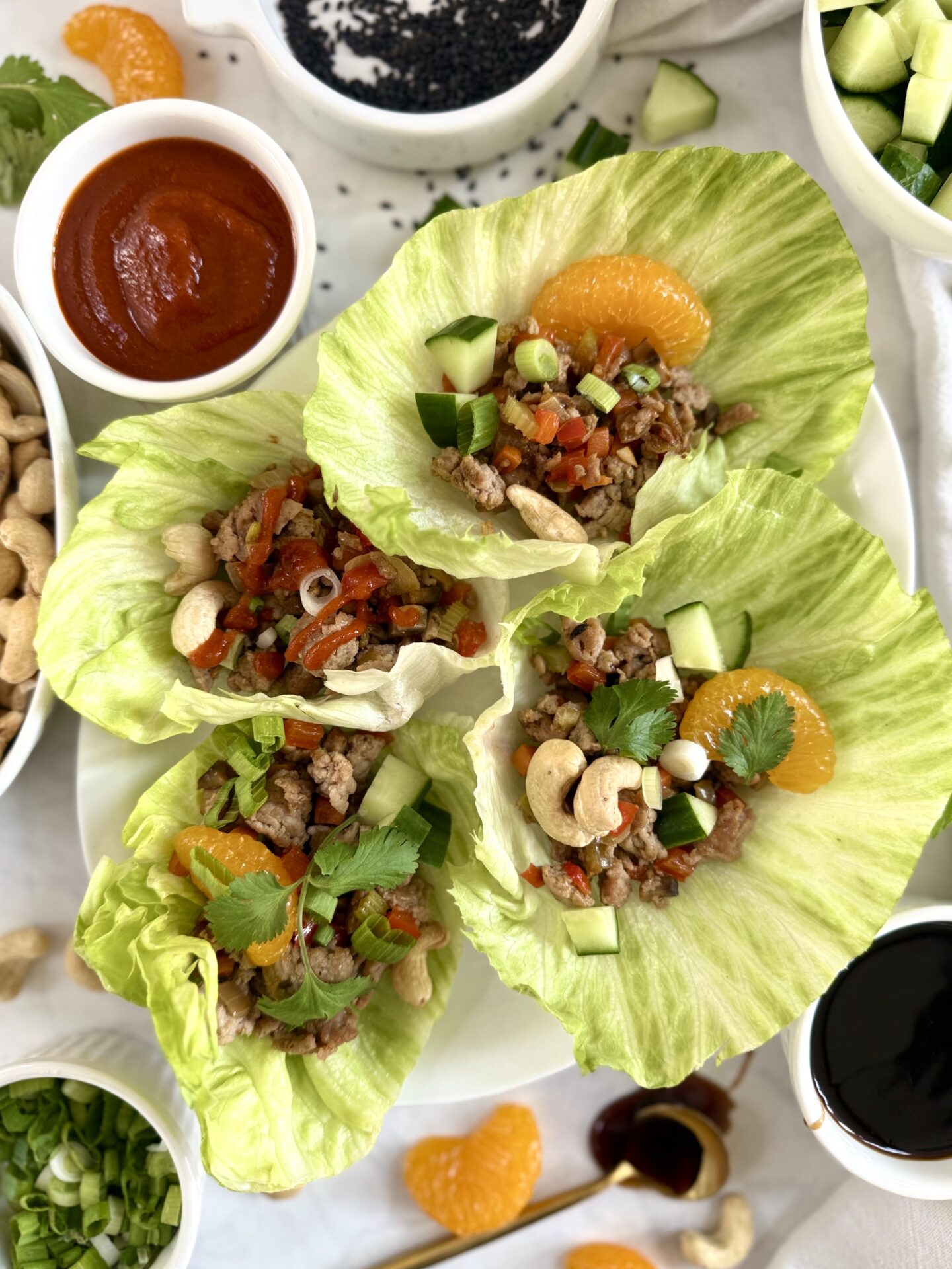 A plate of Ground Turkey Lettuce Wraps is seen from above, surrounded by dishes of chopped cucumber, spring onions, hoisin sauce, cashews and sriracha sauce.