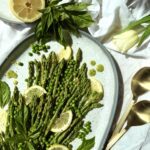 A platter of Lemon Basil Asparagus and Peas is seen from above, lit with bright spring sunshine. Lemons and fresh basil surround the platter that rests on a white table cloth. Pale yellow tulips and gold serving spoons complete the image.