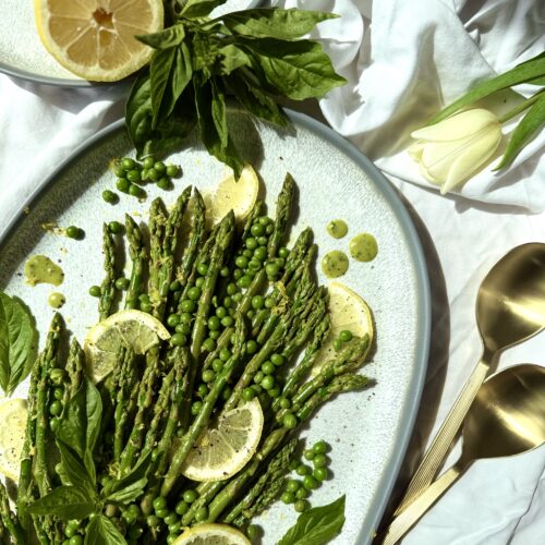 A platter of Lemon Basil Asparagus and Peas is seen from above, lit with bright spring sunshine. Lemons and fresh basil surround the platter that rests on a white table cloth. Pale yellow tulips and gold serving spoons complete the image.