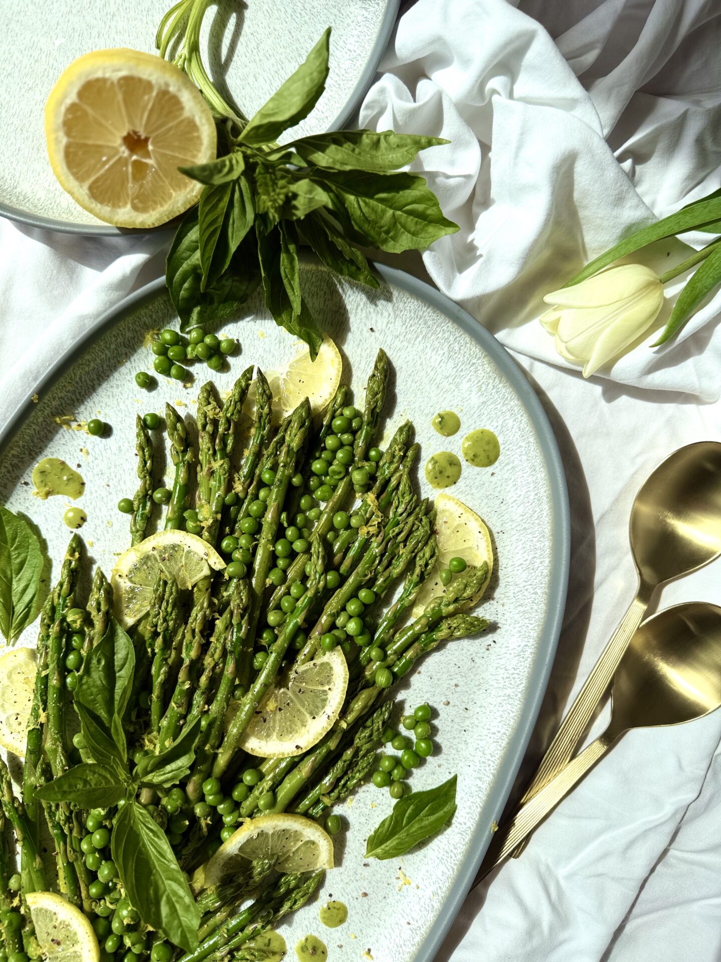 A platter of Lemon Basil Asparagus and Peas is seen from above, lit with bright spring sunshine. Lemons and fresh basil surround the platter that rests on a white table cloth. Pale yellow tulips and gold serving spoons complete the image.