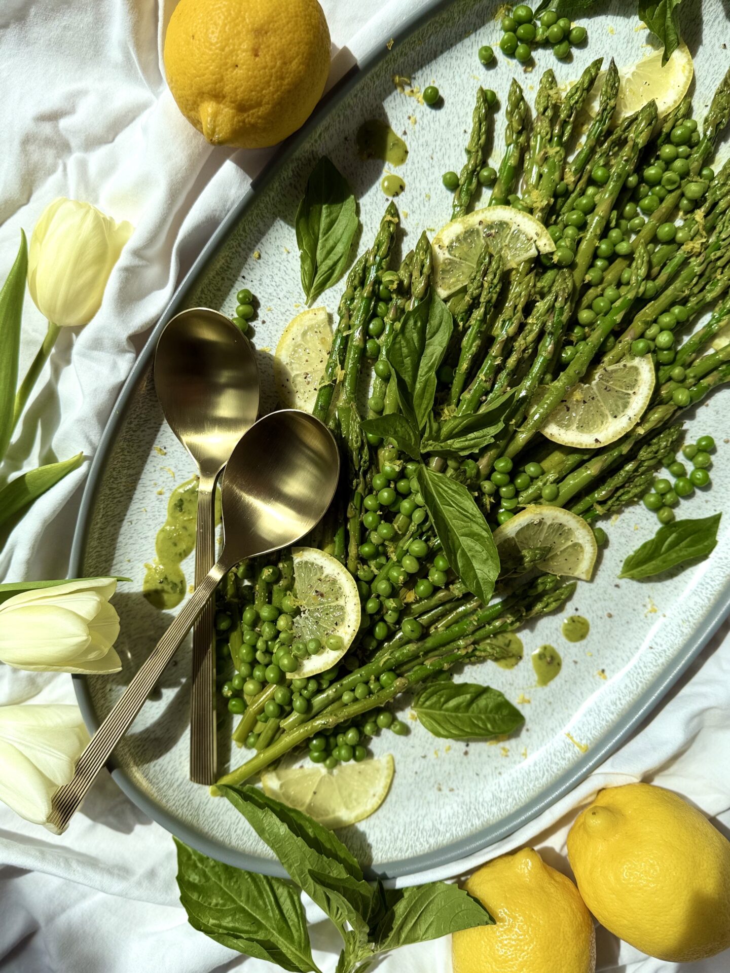 A platter of Lemon Basil Asparagus and Peas is seen from above on a white table cloth.  The vegetables are garnished with lemon wedges, fresh basil leaves, vinaigrette drips and freshly ground black pepper.  A set of gold serving spoons rest on the platter.  Tulips and lemons complete the image.