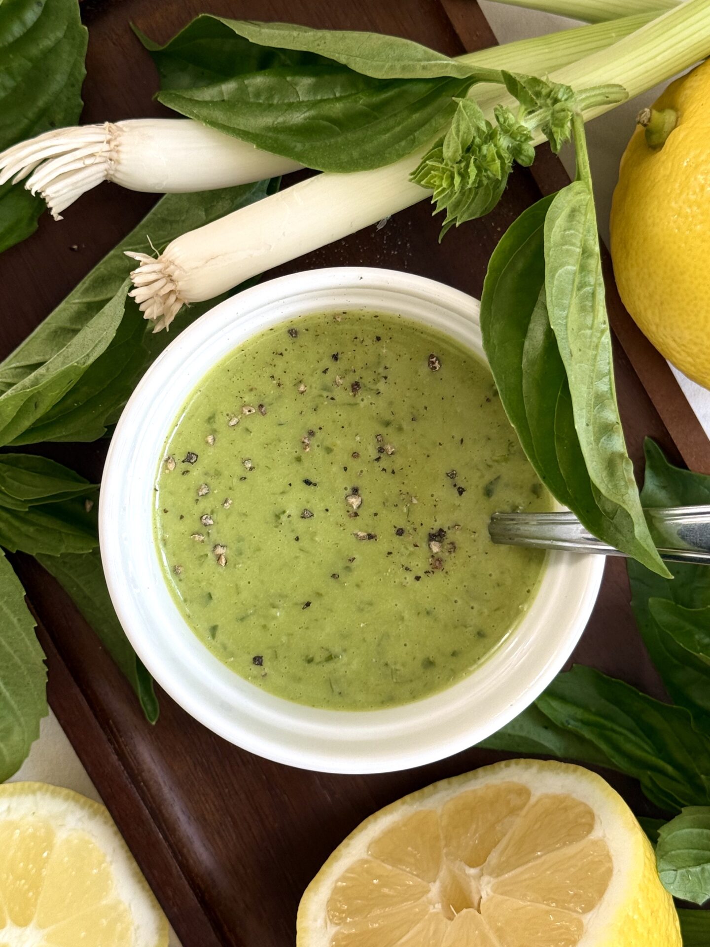 A bowl of Homemade Lemon Basil Dressing sits on a dark wood tray, surrounded by lemons, fresh basil leaves and green onions.