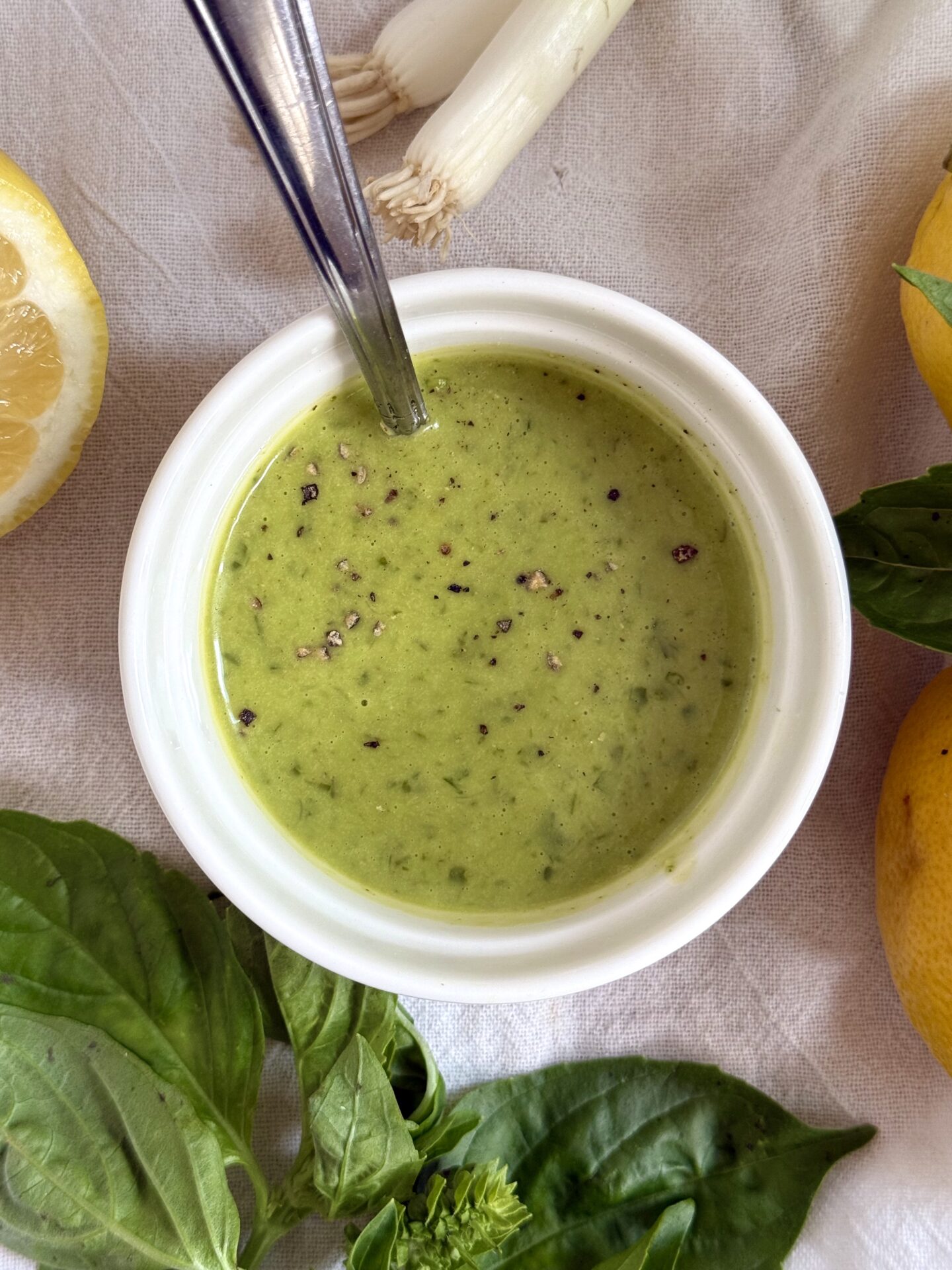 A bowl of Lemon Basil Vinaigrette is seen from above surrounded by fresh basil, lemons and spring onions.