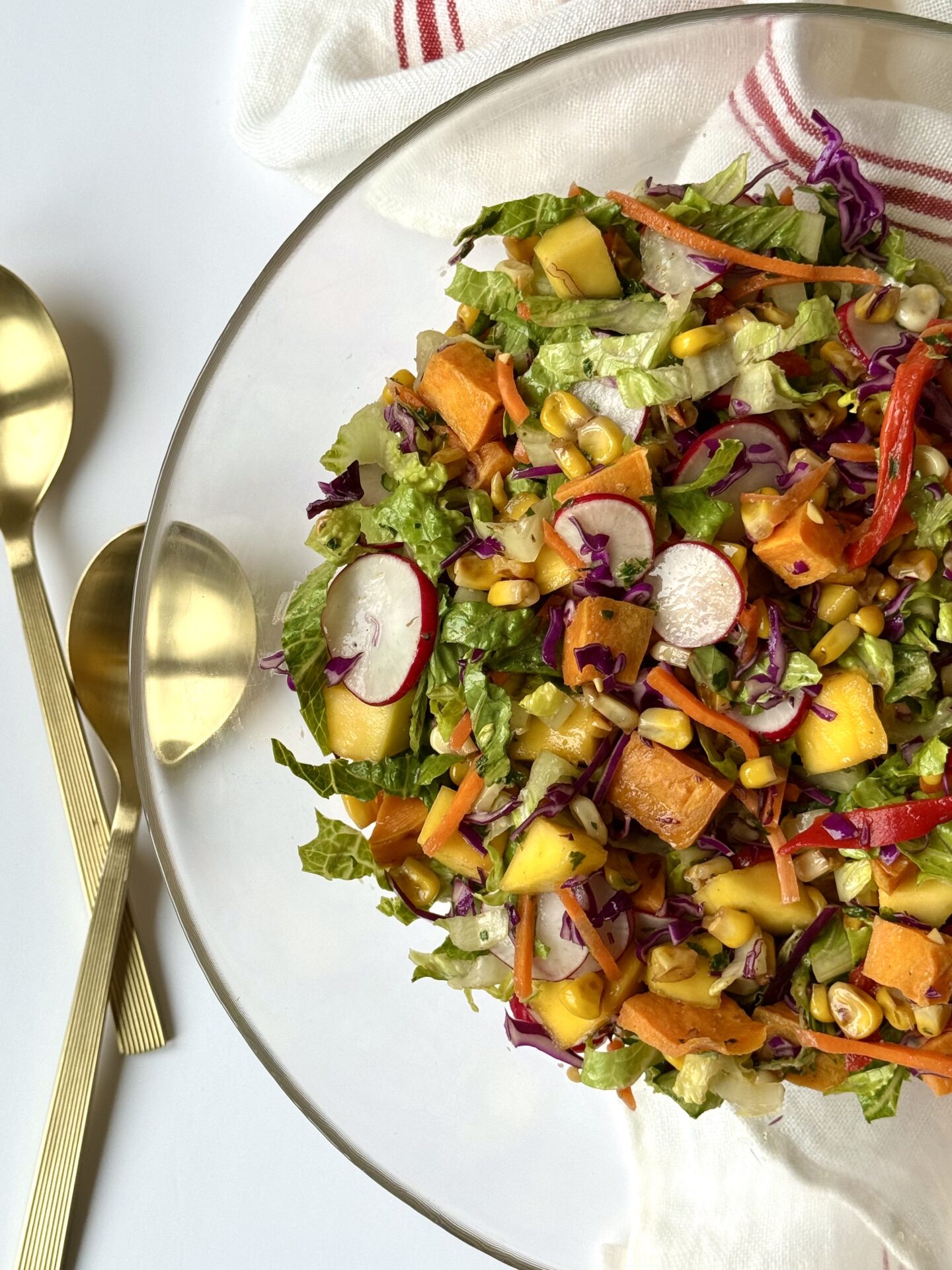 A large glass serving bowl filled with colourful Mexican Chopped Salad is seen from above, with gold coloured salad servers seen alongside.