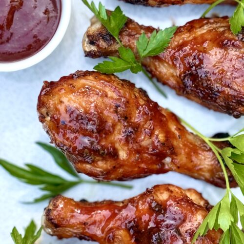 Smokey Sweet Grilled Chicken Legs are seen from above, on a platter, garnished with a small dish of barbecue sauce and a sprinkling of fresh parsley.