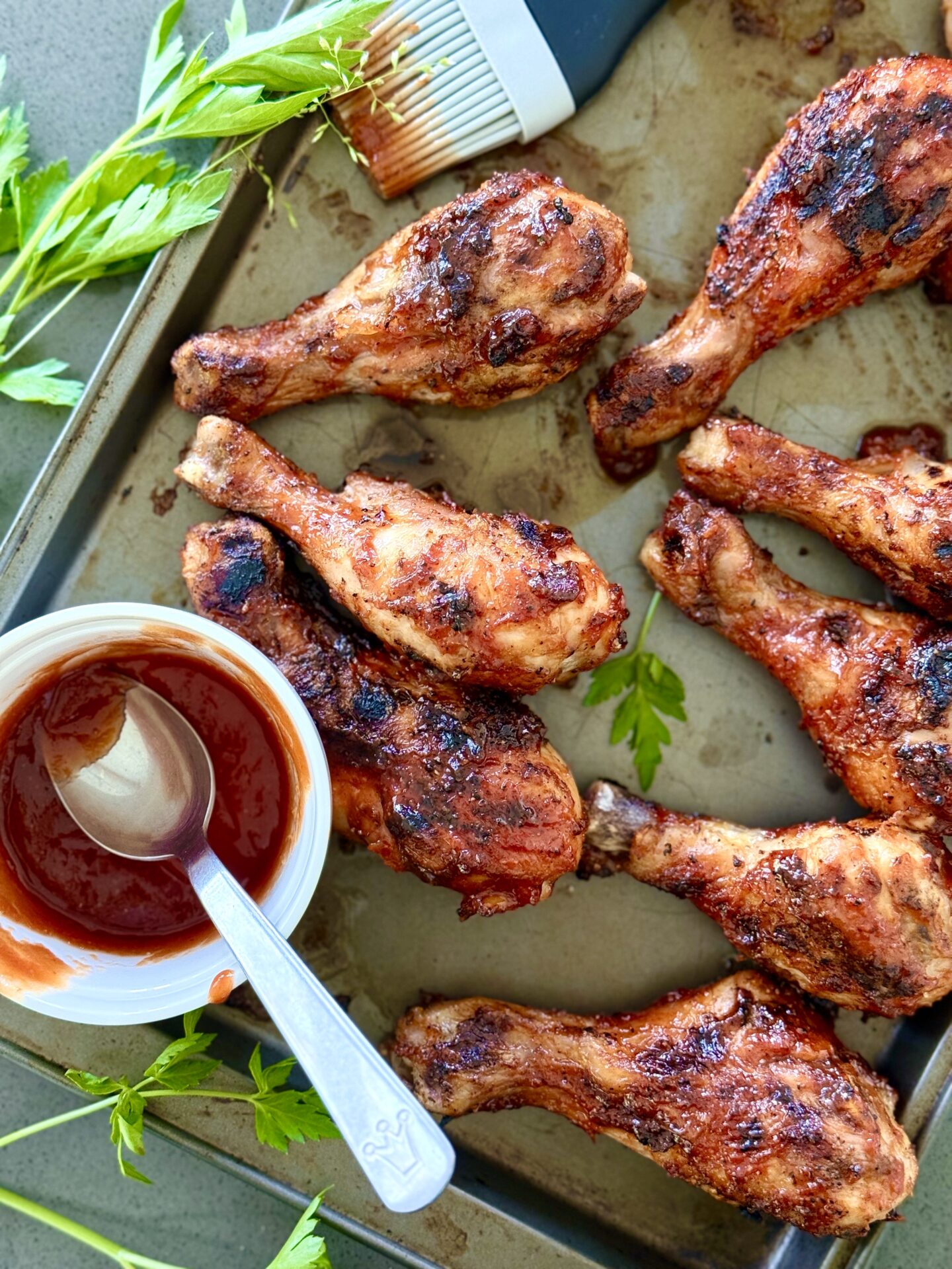 A baking sheet is filled with Smokey Sweet Grilled Chicken Legs. A bowl of homemade barbecue sauce and a basting brush are seen on the edges.
