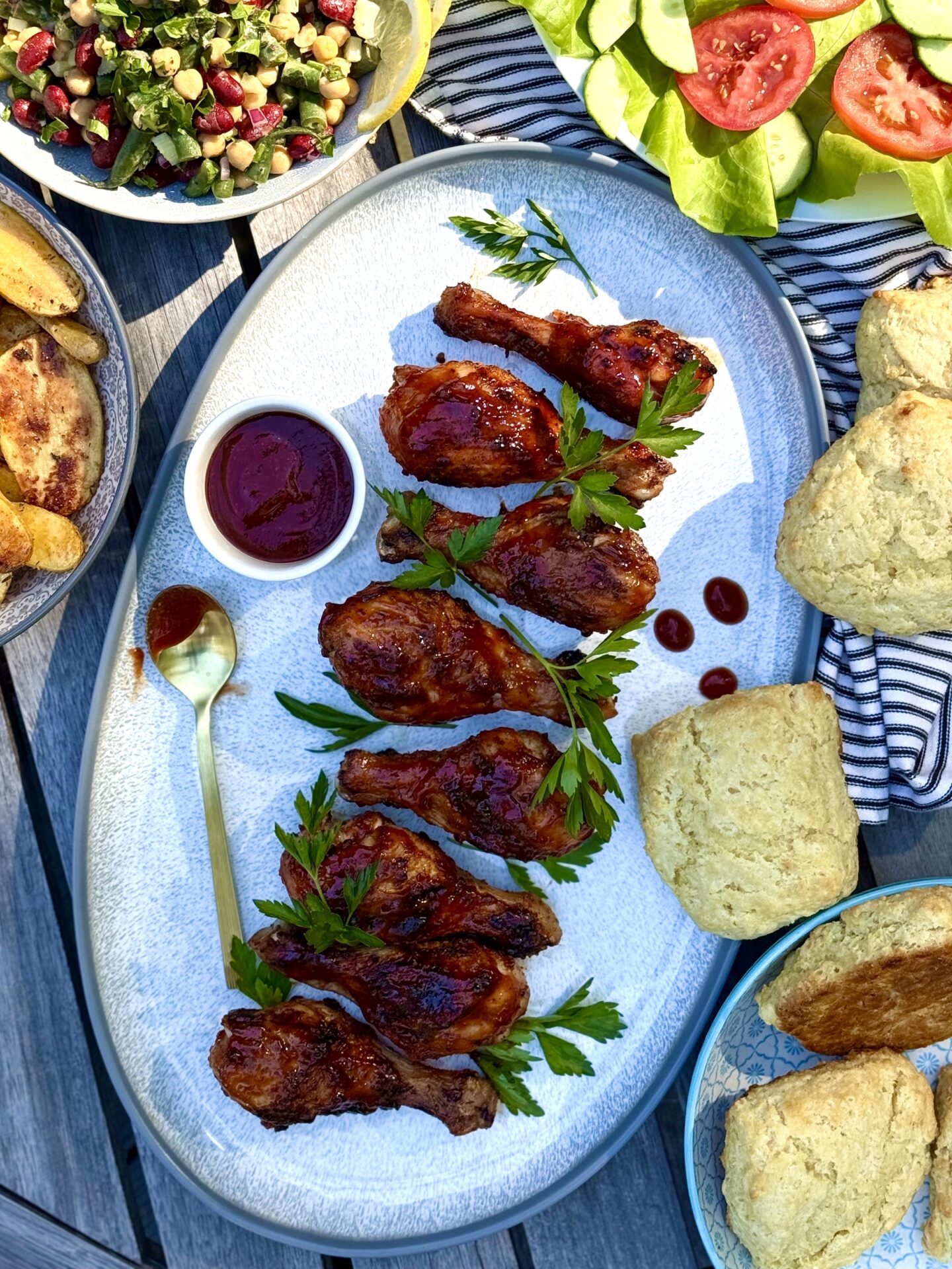 Smokey Sweet Gilled Chicken Legs are lined up on a platter and set on an outdoor dining table.  Plates and bowls of summer side dishes surround the chicken legs.