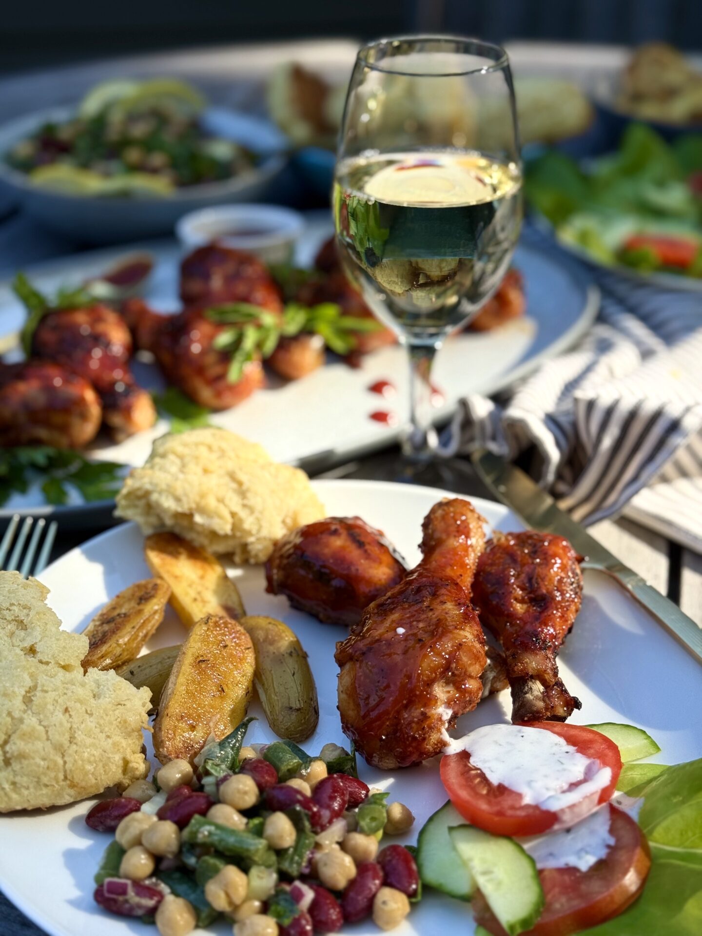An outdoor table filled with platters of Smokey Sweet Grilled Chicken Legs, bean salad and other side dishes is seen in the fading evening sunlight. A glass of white wine sits in the centre of the table catching the light.