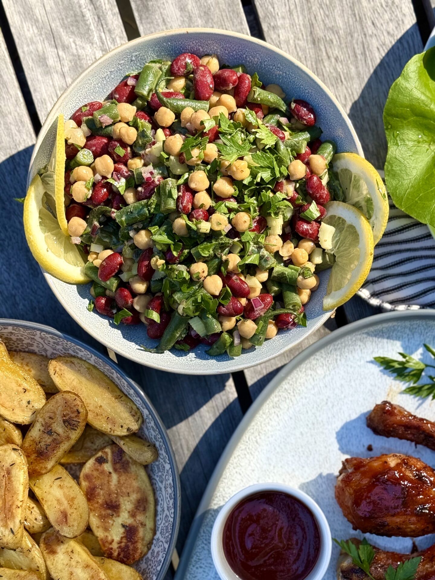 A bowl of Marinated Mediterranean Bean Salad is seen from above on an outdoor table, as part of a backyard barbecue dinner.  The salad is surrounded by platters of grilled chicken and roasted potatoes. 