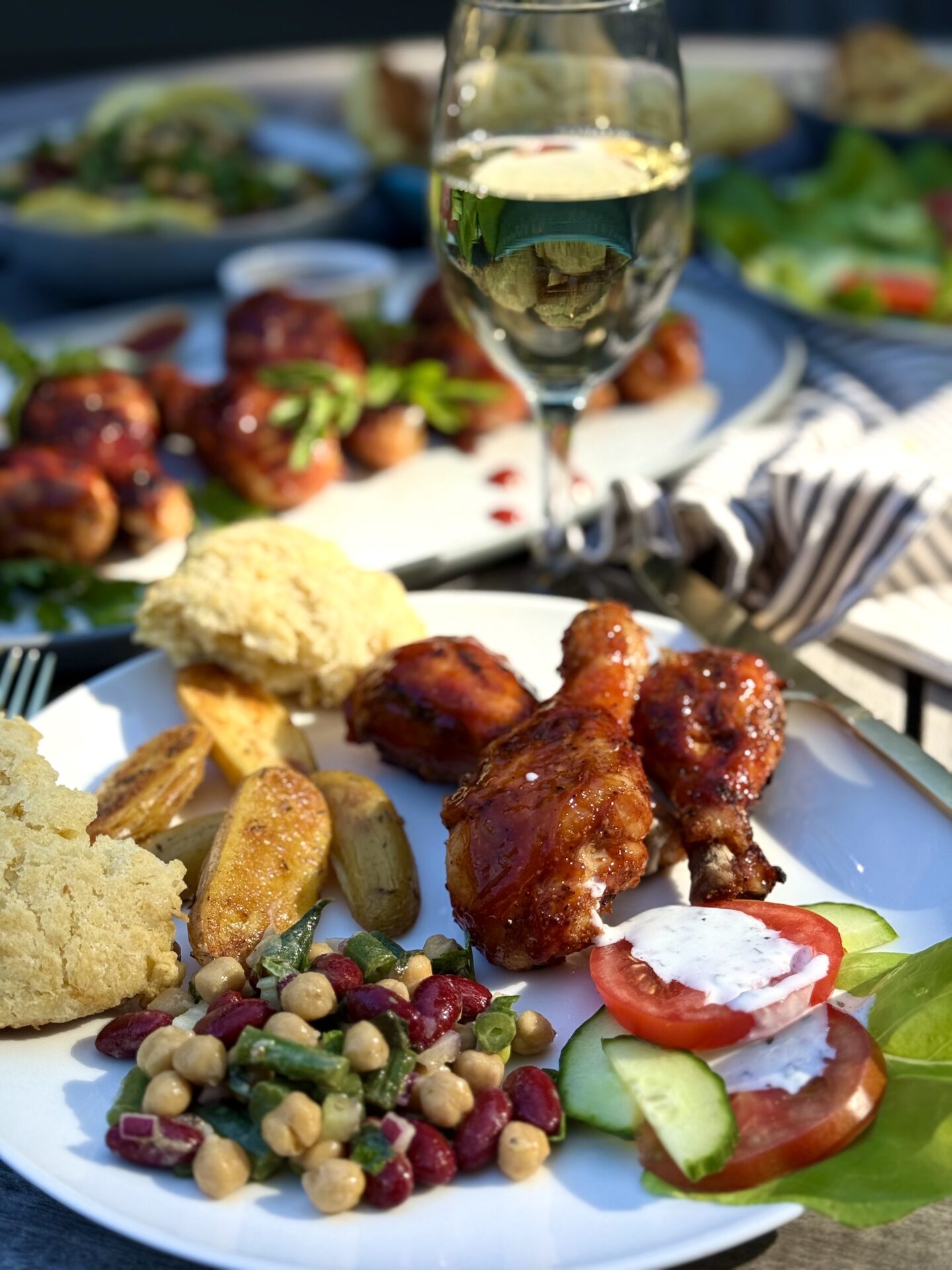 A dinner plate of Marinated Mediterranean Bean Salad with roasted potatoes, salad and grilled chicken is seen on a picnic table.  A glass of cold white wine and platters of food are seen in the background.
