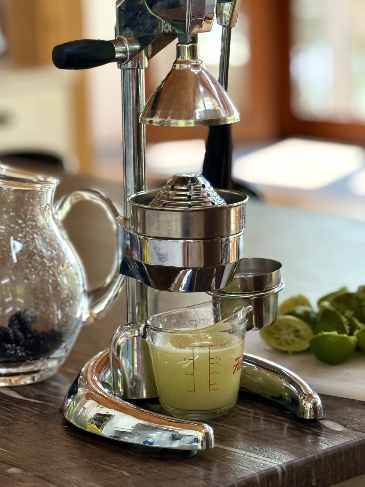 A citrus press sits on a grey stone countertop where it has been pressing fresh limes. A pitcher full of lime juice sits under the press and sunshine streams in through a screen door in the background.
