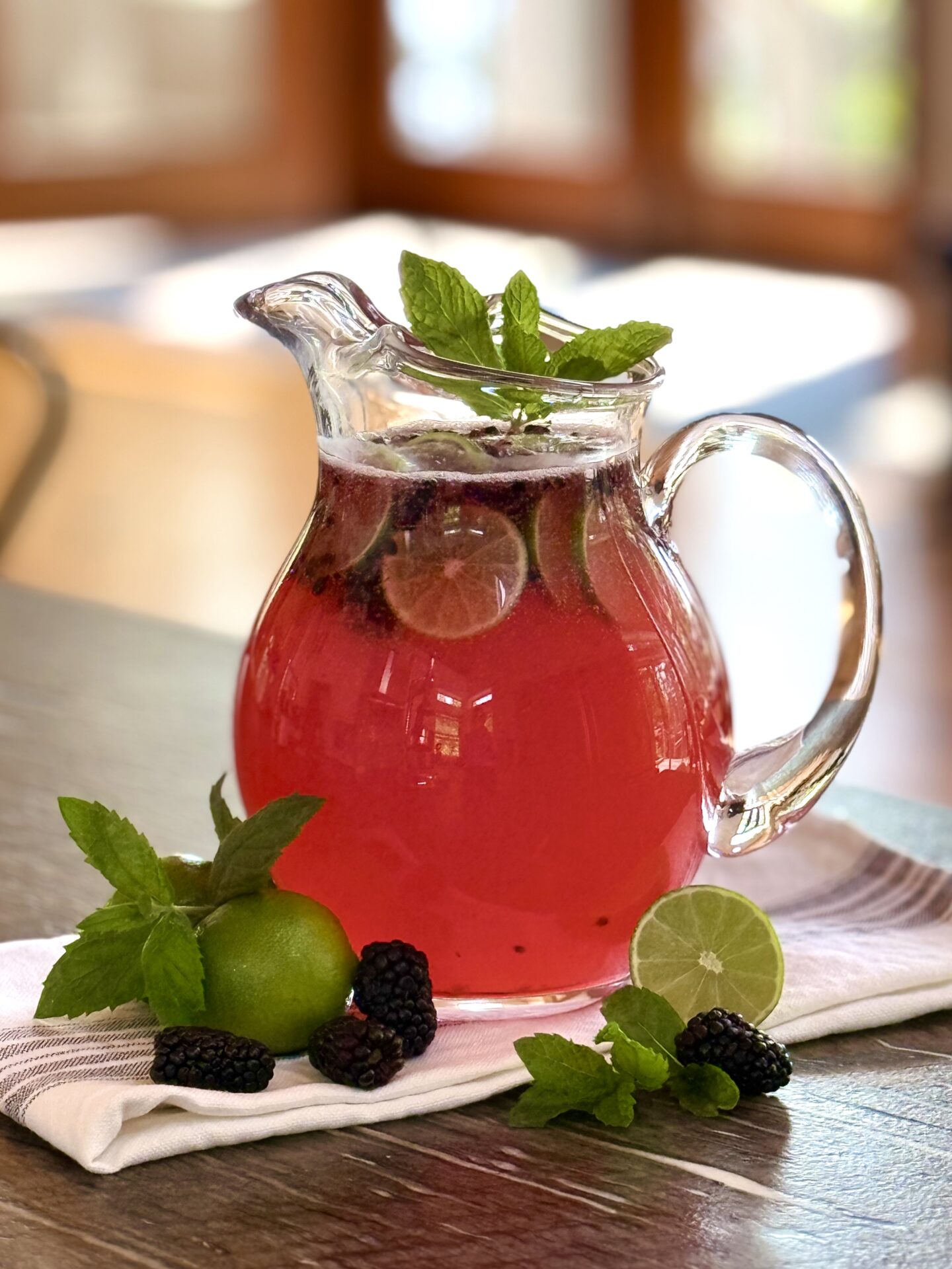 A glass pitcher full of Blackberry Mojito Mocktail sits on a table, garnished with blackberries, lime wedges and fresh mint. Sunshine streams in through the screen doors in the background. 