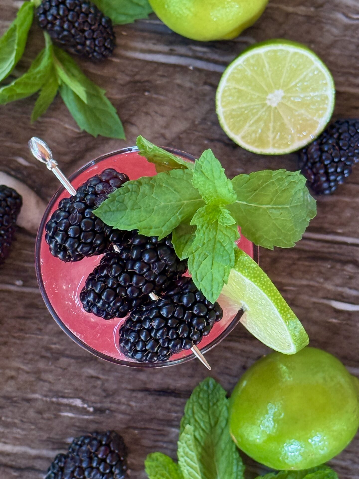 A glass of Blackberry Mojito Mocktail is seen from above garnished with a cocktail pick strung with plump blackberries, a lime wheel and sprigs of fresh mint.