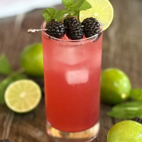 A tall glass of Blackberry Mojito Mocktail sits on a grey stone countertop , garnished with plump blackberries, lime wheels and fresh mint sprigs.