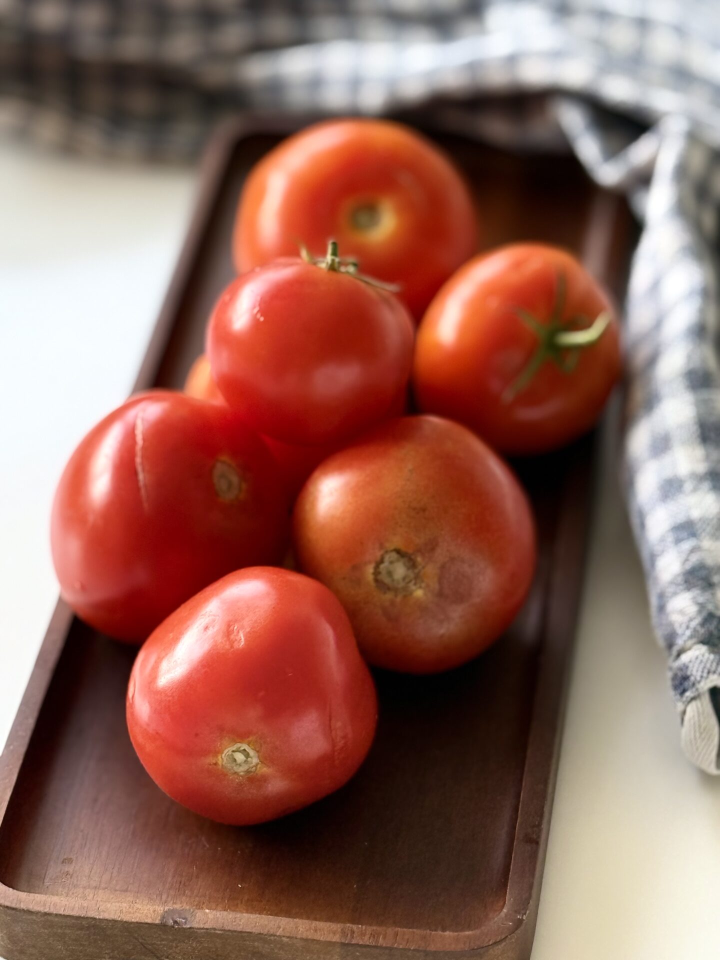 A wood tray is stacked high with garden fresh tomatoes of varying sizes. A checkered cloth is seen in the background. 