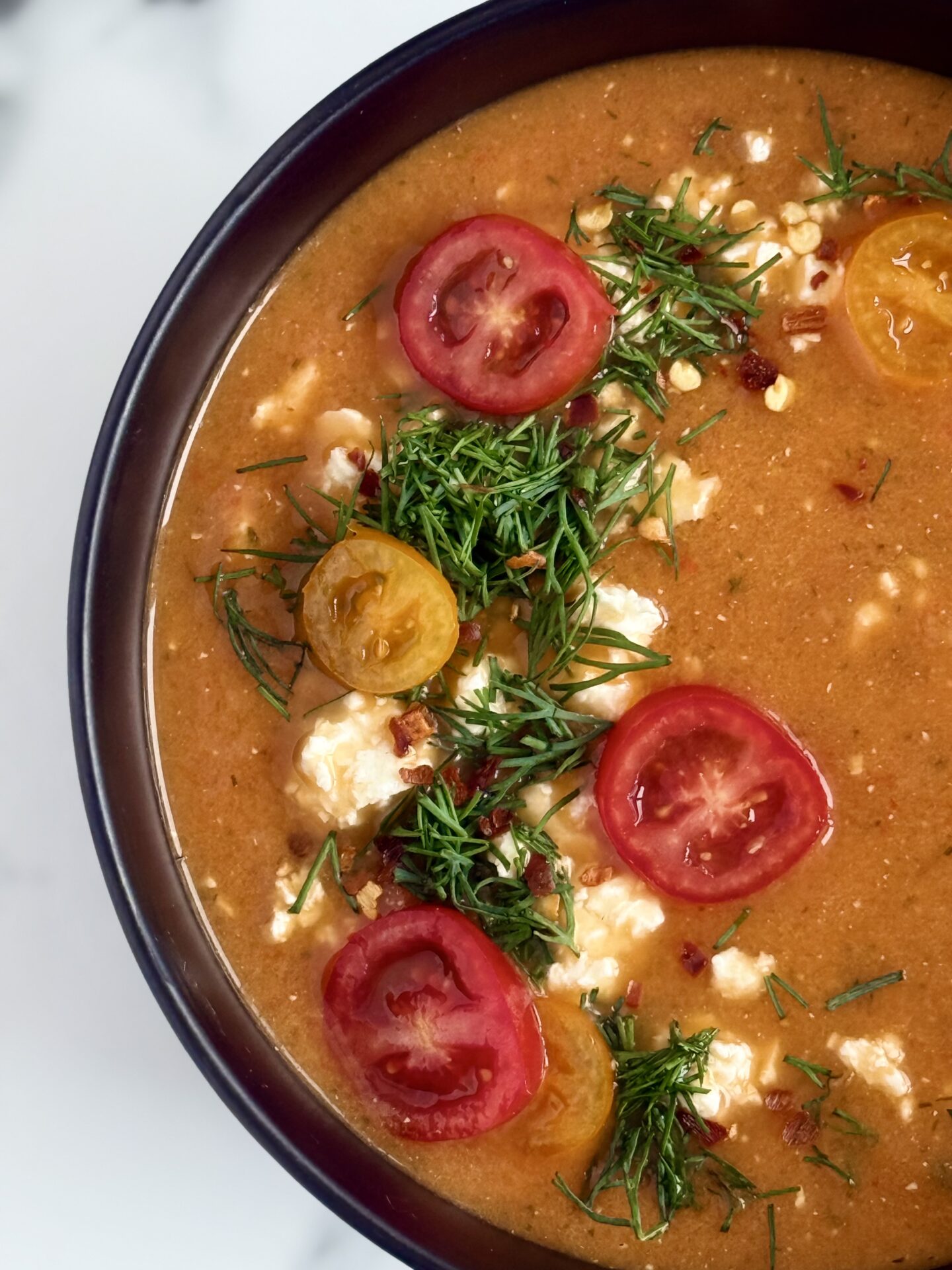 Roasted Tomato and Feta soup is seen close up, to focus on the garnish of fresh dill, crumbled feta and sliced cherry tomatoes.
