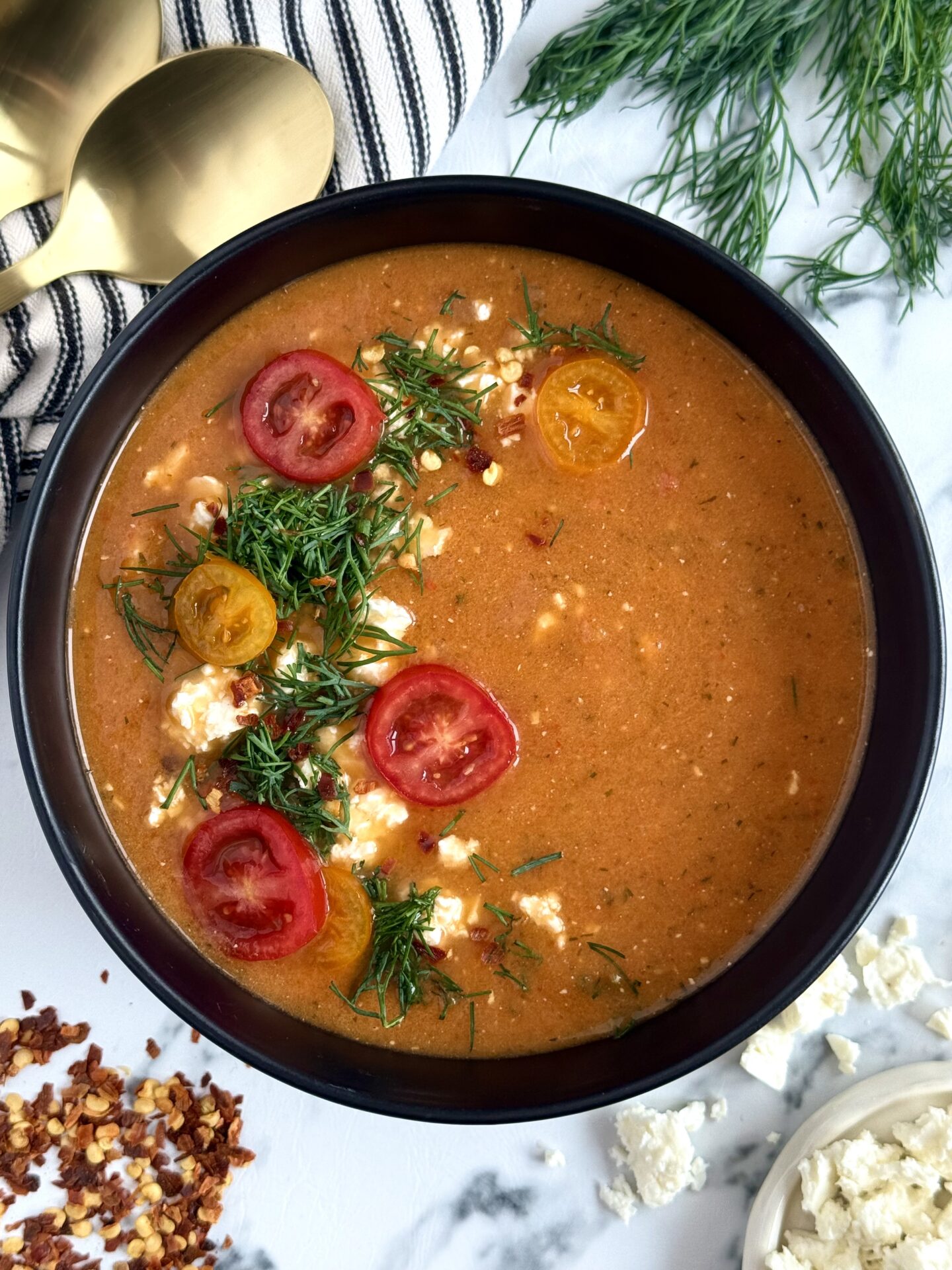 A black bowl filled with roasted tomato and feta soup is seen from above. The soup is garnished with a semi-circle of fresh chopped dill, crumbled feta cheese and sliced cherry tomatoes. Fresh dill, feta, chili flakes and gold spoons surround the bowl.