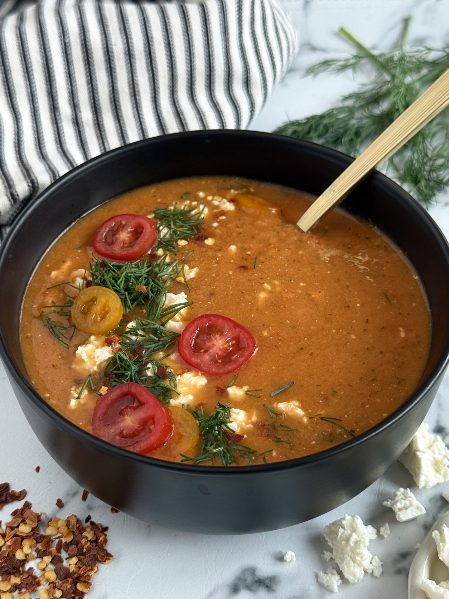 A black bowl filled with Roasted Tomato and Feta soup sits on a marble counter, surrounded by a black and white striped cloth, chili flakes, fresh dill and crumbled feta. A gold spoons surround sits in the soup, that is beautifully garnished.