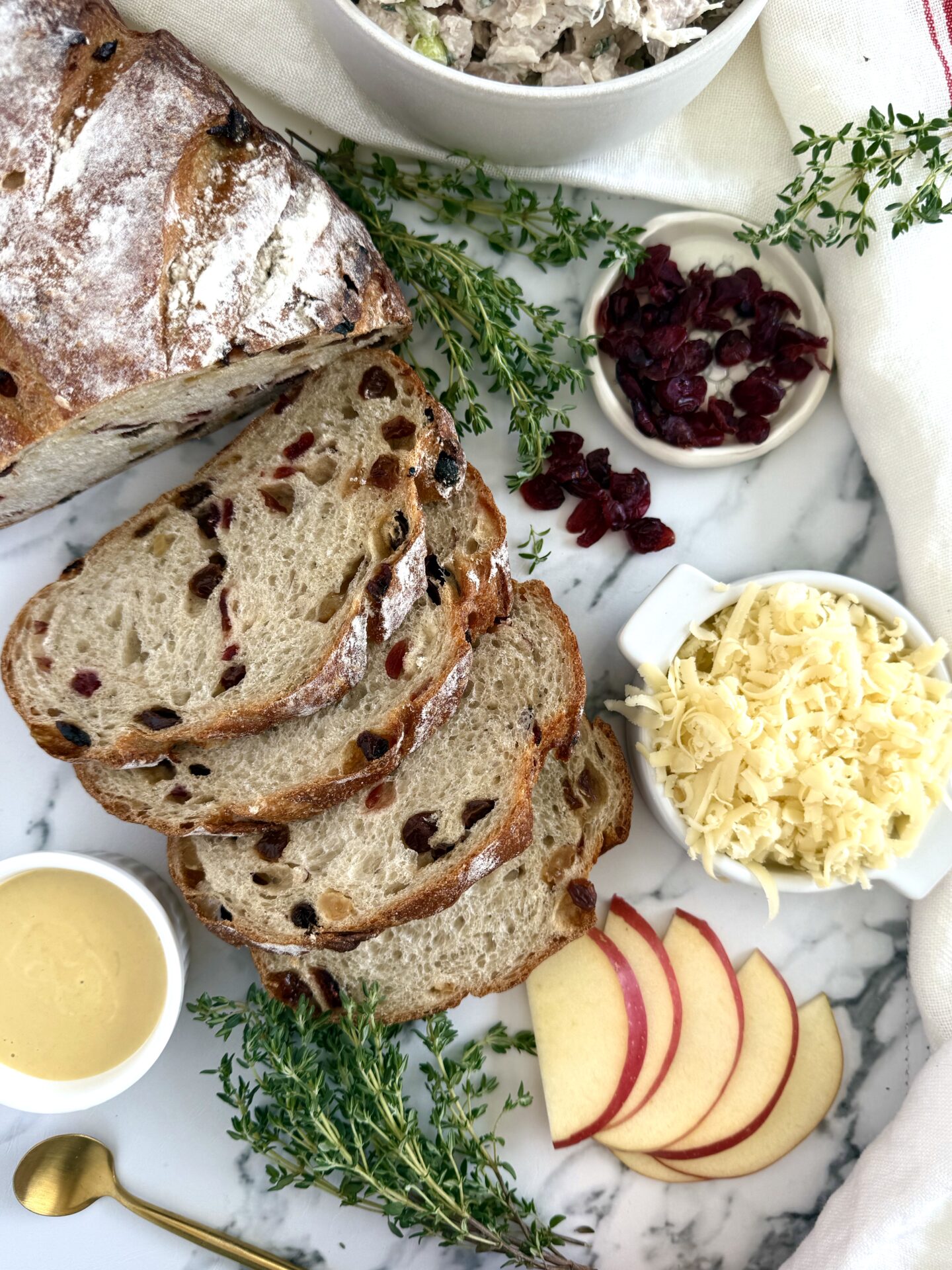 The ingredients for Chicken and Apple Melt Sandwiches with Smoked Cheddar are seen from above, resulting on a white marble countertop. A loaf and slices of cranberry-studded bread are surrounded by apple slices, maple mustard, grated smoked cheddar and fresh thyme.