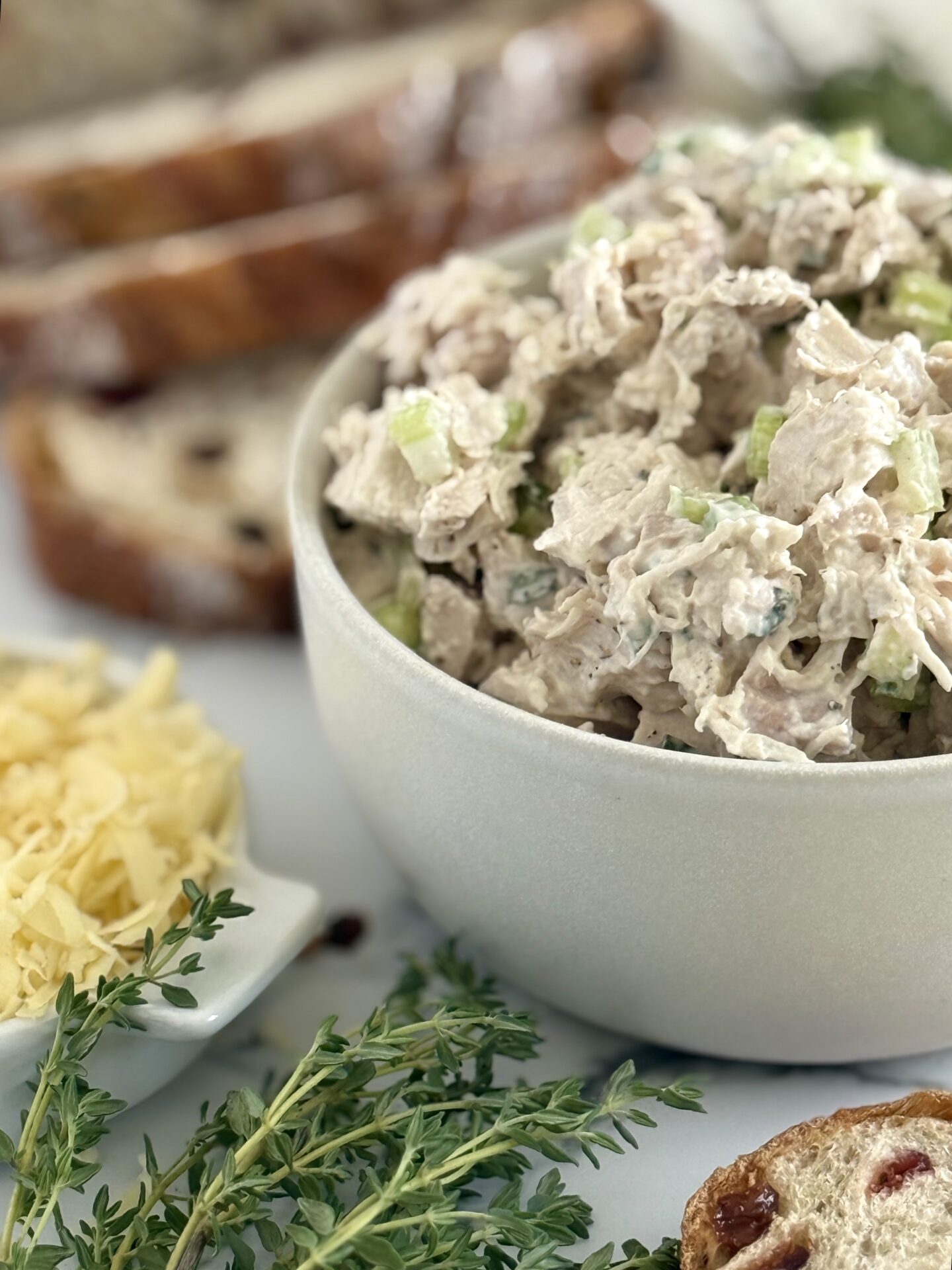 A bowl of creamy chicken salad is seen beside a dish of grated smoked cheddar cheese. A stack of cranberry bread islices is seen n the background.