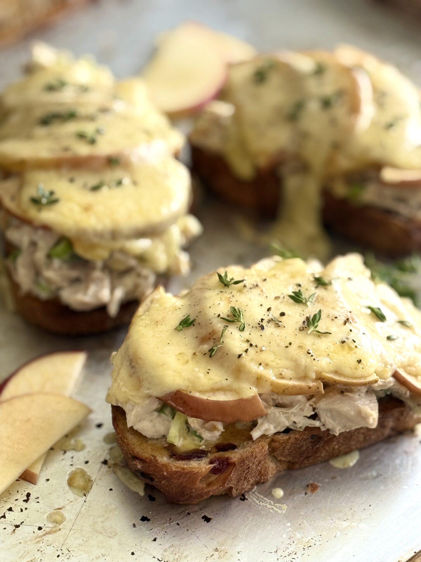Chicken and Apple Melts sit on a metal baking sheet, hot from the oven, with gooey, melted smoked cheddar cheese dripping from the edges of the sandwiches.