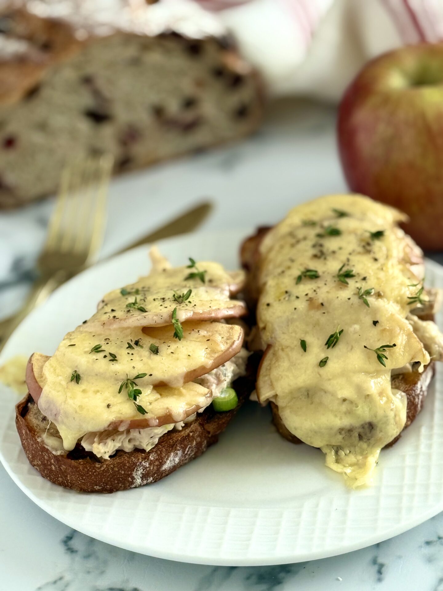 Chicken and Apple Melt open-faced sandwiches are seen on a white plate. The smoked cheddar cheese is melted over the tops and drips down the sides of the sandwiches that are stacked high with chicken salad and apple slices.