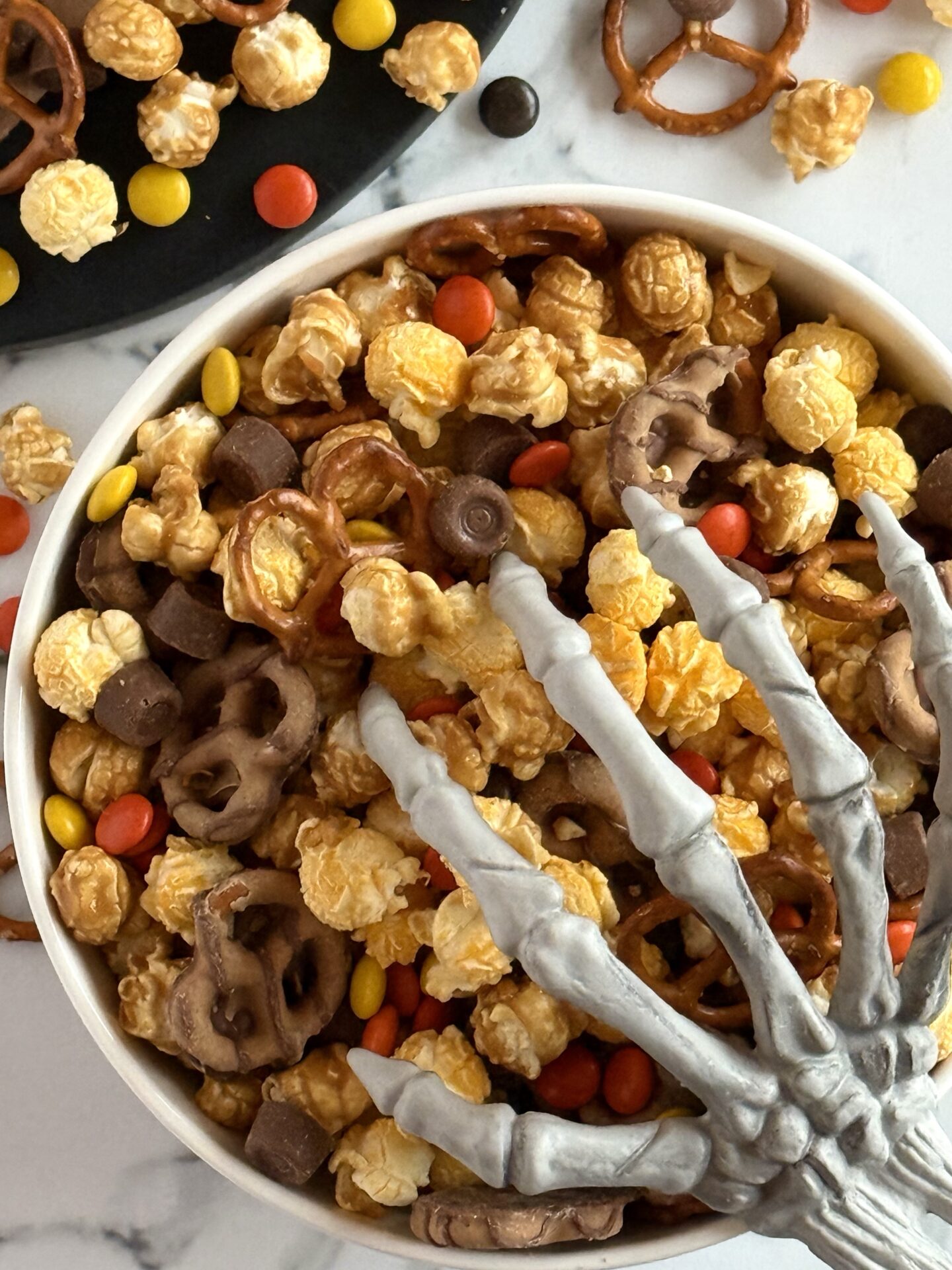 Sweet and salty Monster Munch Halloween Snack Mix is seen spilling over onto the counter.  A bony hand reaches in from the corner.