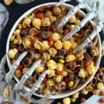 A bowl of Monster Munch Halloween Snack Mix is seen from above. Skeletons hands reach into the bowl from either side.
