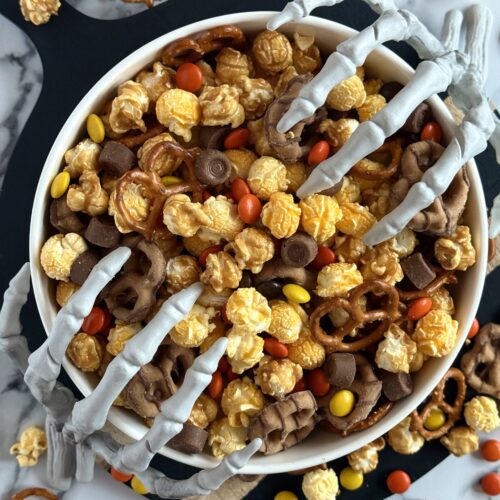 A bowl of Monster Munch Halloween Snack Mix is seen from above. Skeletons hands reach into the bowl from either side.