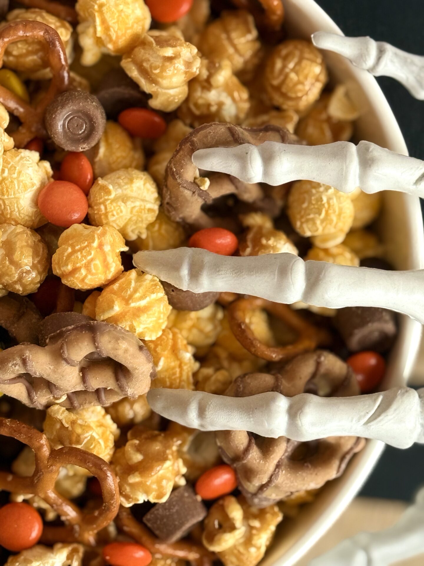 A bowl of Monster Munch Halloween Snack Mix is seen close up with a skeleton hand creeping into the bowl from the side.