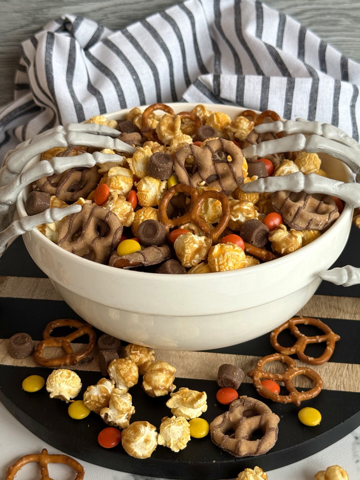 A bowl of Monster Munch Halloween Snack mix spills over onto a black wooden board.  Skeletons hands reach hands creep into the bowl from either side.