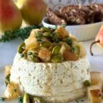 An elegant Pistachio and Pear Baked Ricotta sits in the foreground with chunks of caramelized pear, bright green pistachio and fresh sprigs of thyme tumbling from the top. The appetizer is surrounded by fresh pears and crackers.