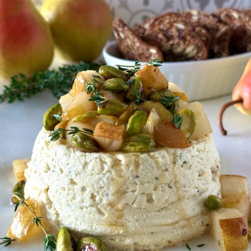 An elegant Pistachio and Pear Baked Ricotta sits in the foreground with chunks of caramelized pear, bright green pistachio and fresh sprigs of thyme tumbling from the top. The appetizer is surrounded by fresh pears and crackers.