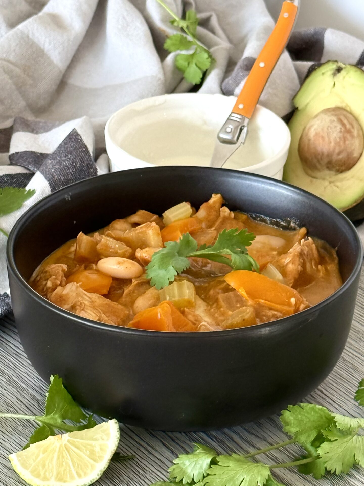 A black bowl of Pumpkin Chicken Chili sits on a table, surrounded by fresh avocado, cilantro, lime wedges and a dish of sour cream.