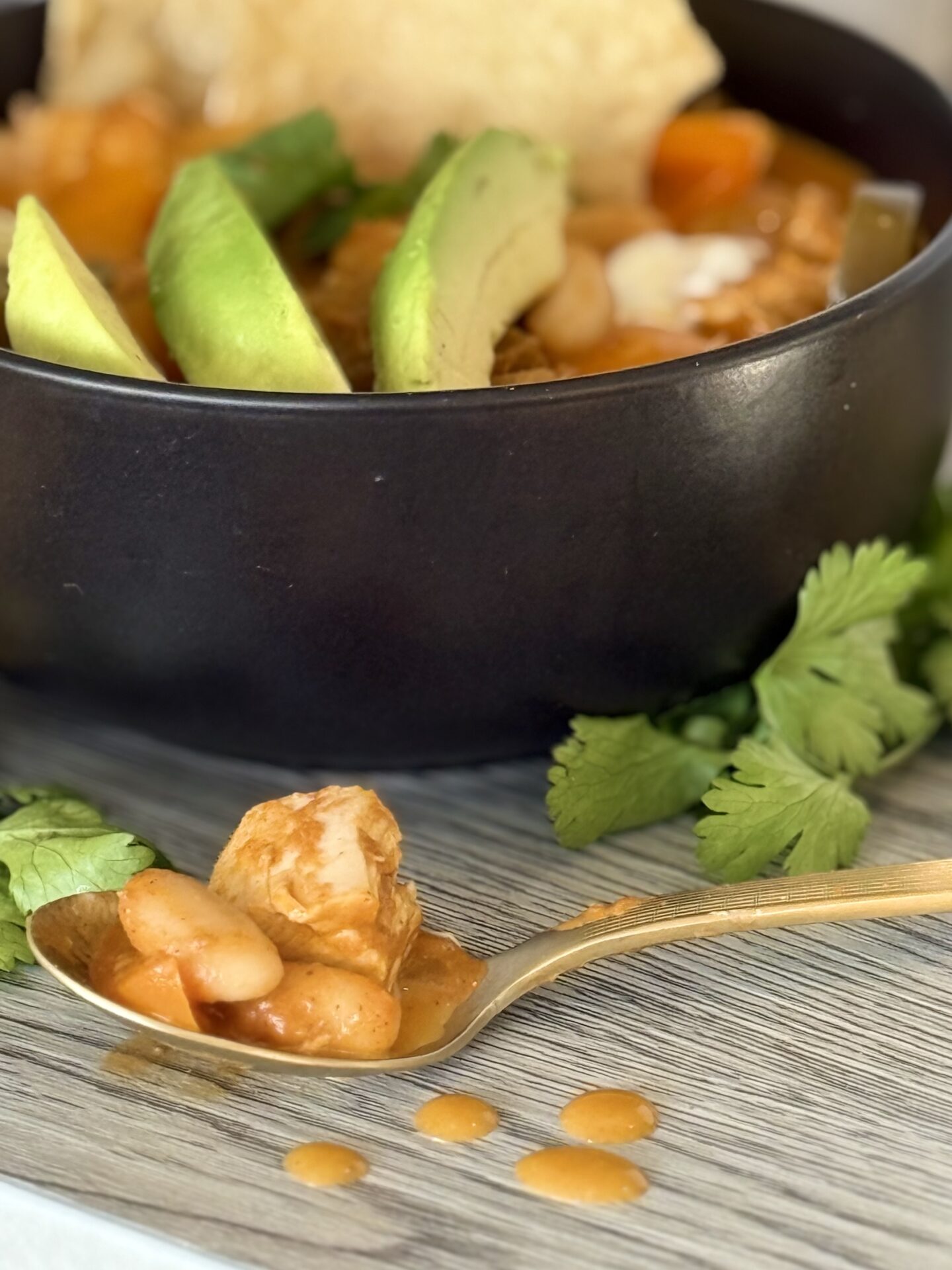 A spoonful of Pumpkin Chicken Chili rests on a table, surrounded by drips of its delicious orange hued sauce. A black bowl full of lavishly garnished chili sits in the background, overflowing with fresh avocado, cilantro and corn chips.