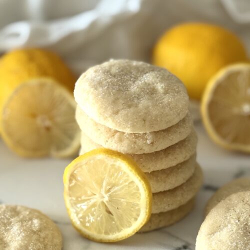 A stack of glittering chewy lemon sugar cookies is seen on a white marble countertop, surrounded by whole lemons and fresh lemon slices.
