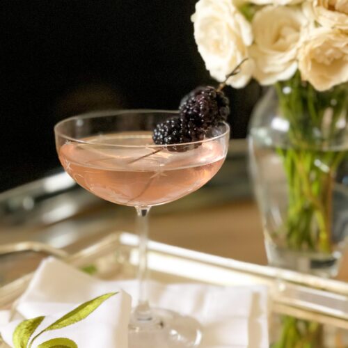 A pretty pink Blackberry Blush Cocktail is served in an elegant coupe glass on a silver tray. A vase of white roses appears in the background.