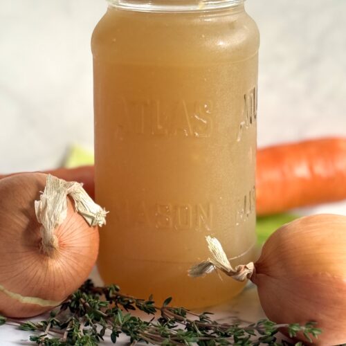 A glass jar of homemade chicken stock sits on a marble counter, surrounded by carrots, celery, onions and fresh thyme.