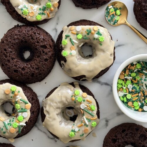 A group of baked chocolate donuts is seen from above. Some are topped with a Baileys glaze and St Patrick’s Day themed sprinkles.